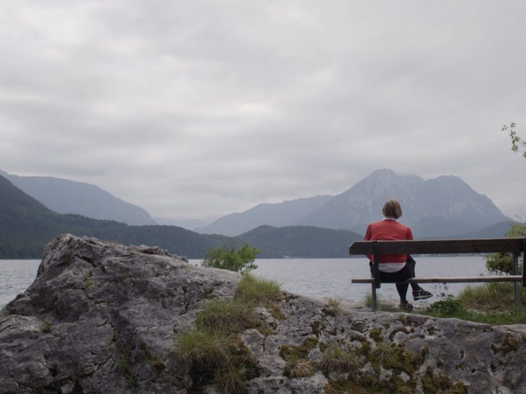 Renate Welsh, Brille, eine rote Halskette, steht in einem grünen, saftigen Garten, die tiefstehende Sonne scheint ihr auf die Haare