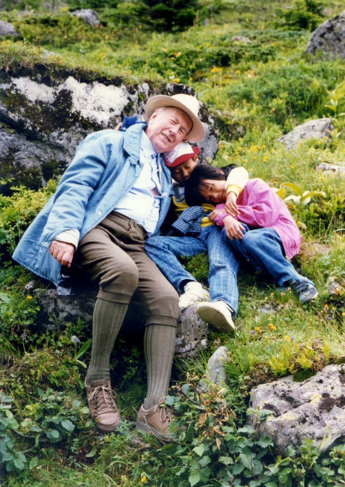Kardinal König in Wanderkleidung sitzt auf einer steilen Bergwiese mit Felsen mit zwei vietnamesischen Kindern, sie kuscheln und lachen