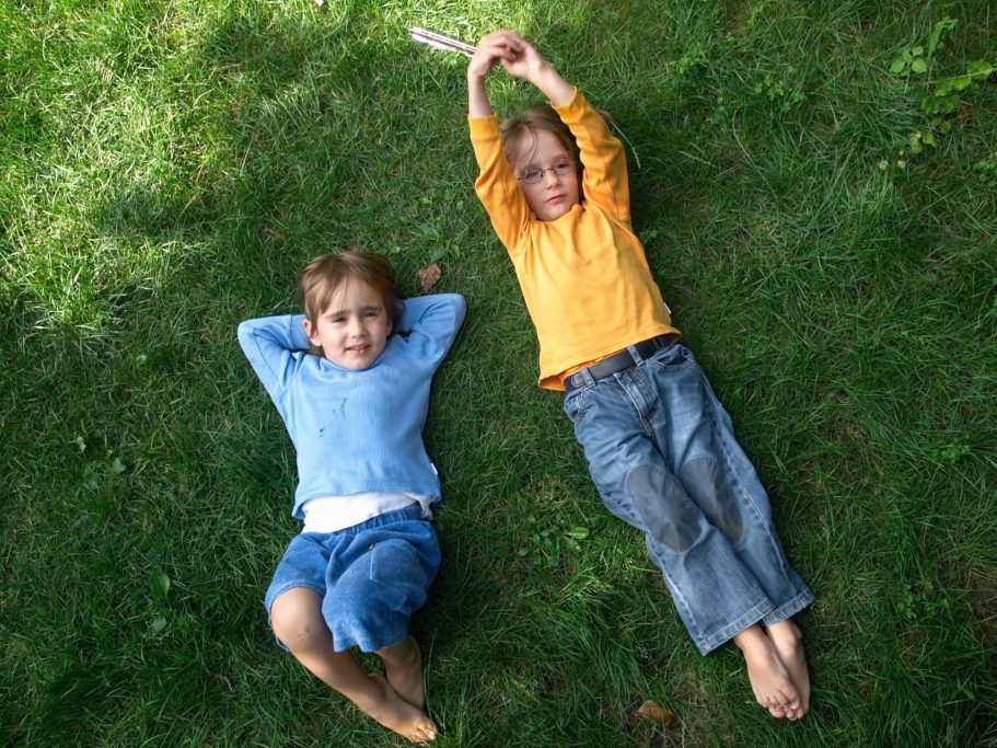 Zwei Kinder liegen entspannt auf grünem Gras, eines in blauem und das andere in orangefarbenem Pullover.