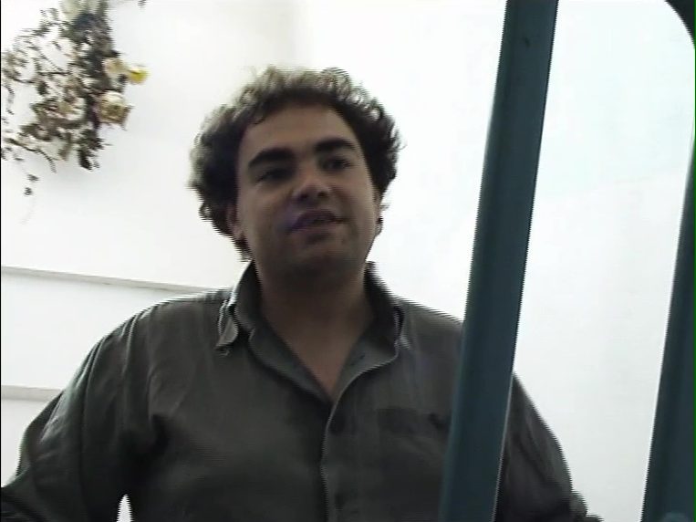 Mann mit lockigem Haar, der vor einer hellen Wand steht und aussieht, als ob er spricht.