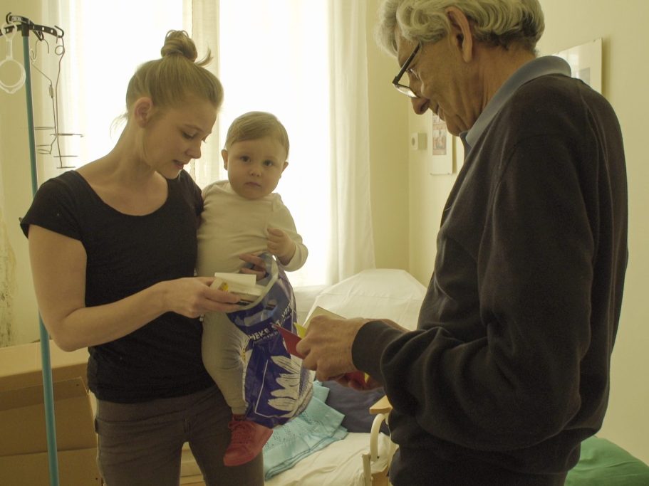 Eine junge Mutter hält ihr Baby im Arm, im anderen Arm ein Sackerl mit Medikamenten, rechts steht Dr. Shiraz Rabady mit dem Mutter-Kind-Pass