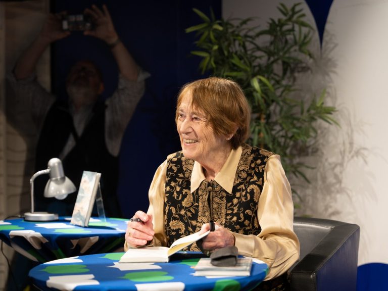 Renate Welsh lächelt, ein offenes Buch vor ihr und hält einen Stift, sie signiert das Buch "Ich ohne Worte"