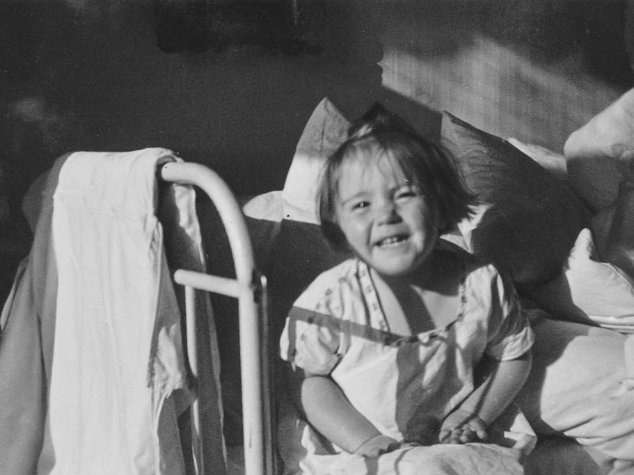 schwarz weißes altes Foto von Renate Welsh als Kleinkind, das in einem Bett sitzt und lacht